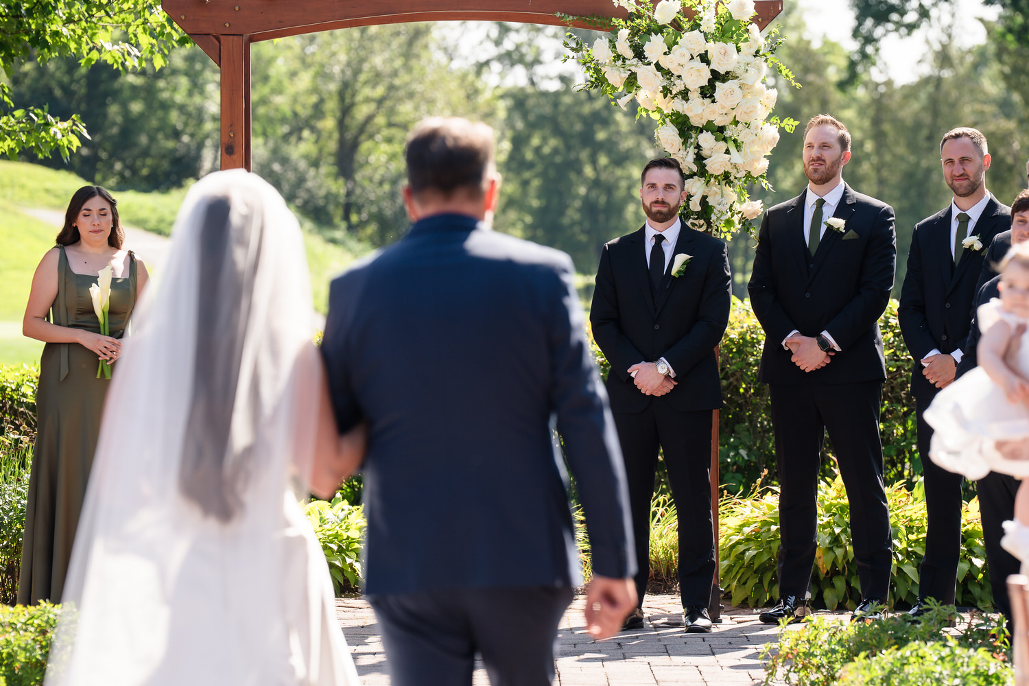 Brude walking toward the outdoor ceremony altar under a wooden arch.
