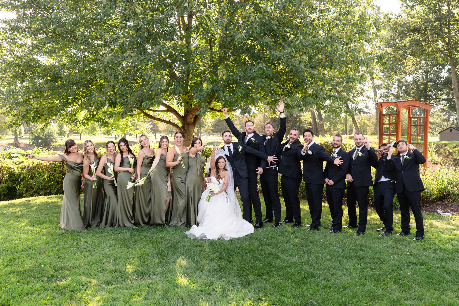 Full wedding party in black suits and sage dresses posing on the green.