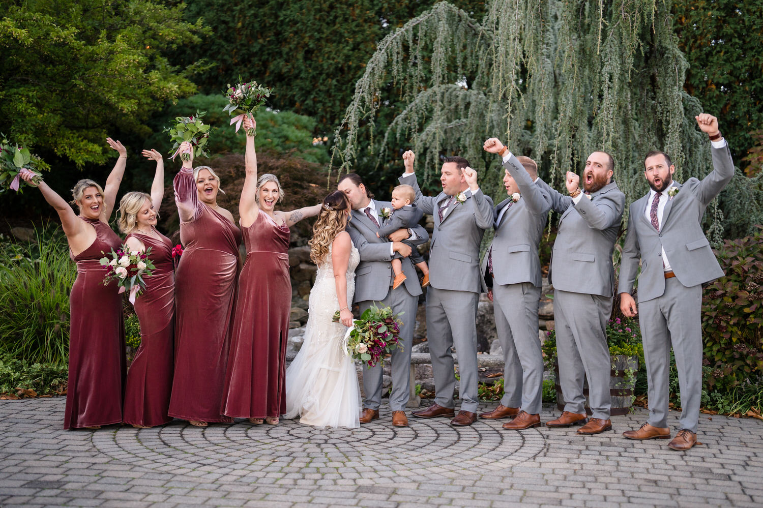 Joyful wedding party celebration shot in the Woodstone Gardens with bridesmaids cheering in burgundy dresses.