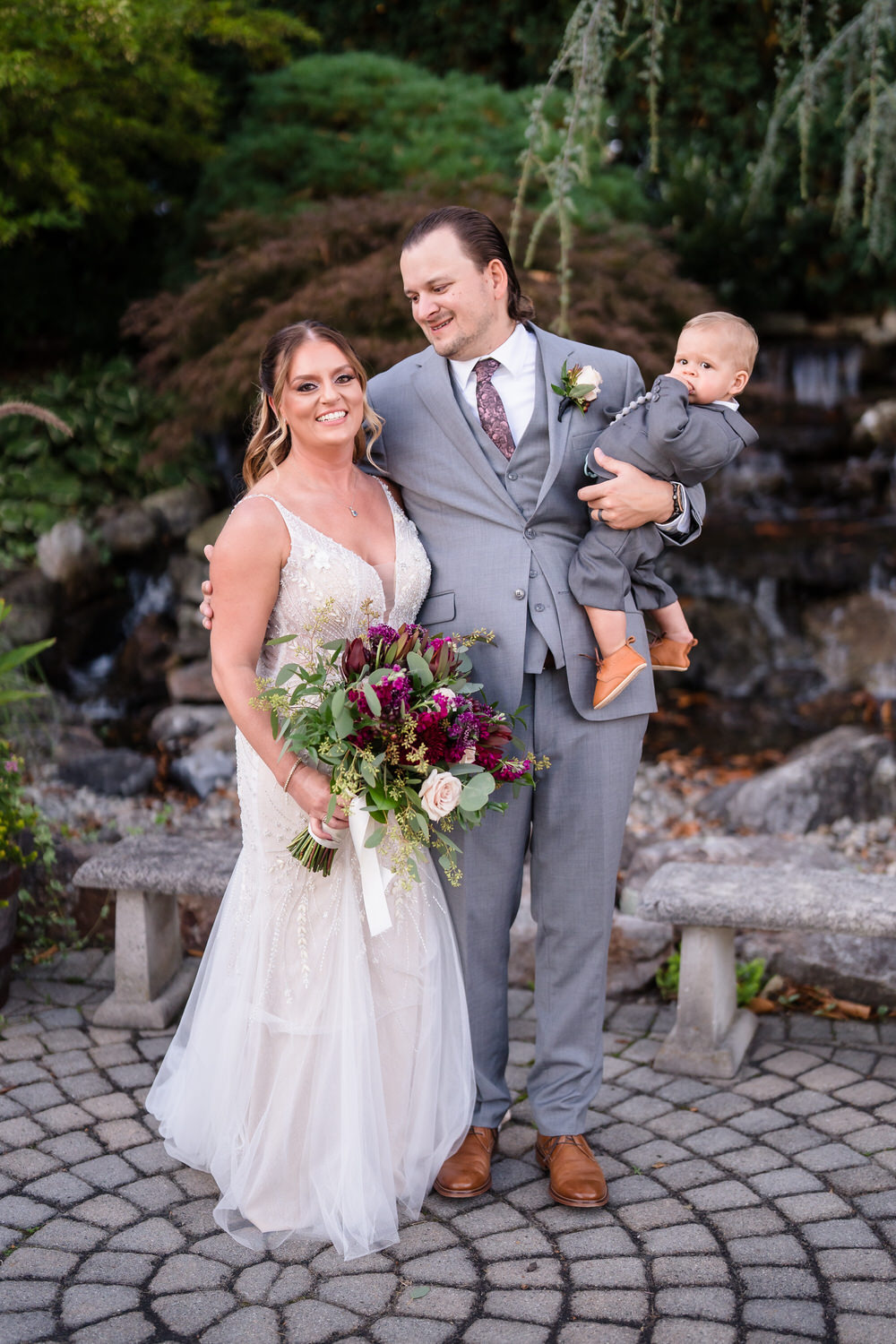 Lifestyle portrait of a bride and groom posing with their young child at their Woodstone Country Club wedding.