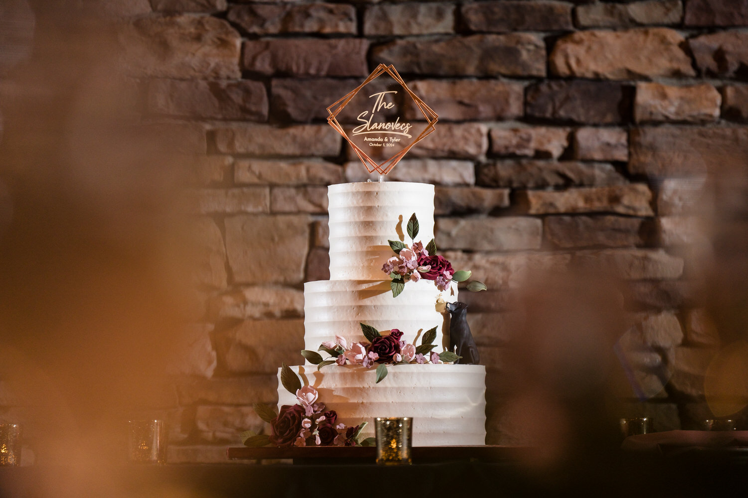 Textured landscape shot of a three-tier wedding cake set against the rustic stone walls of the Woodstone Lodge.