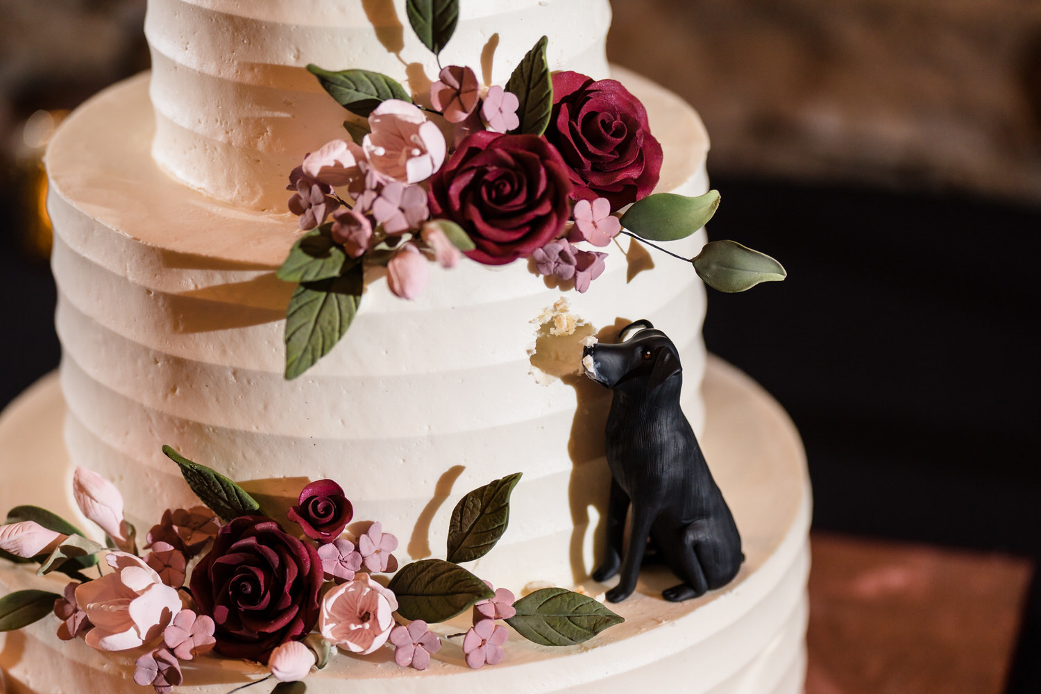 Close-up detail of a three-tier white wedding cake decorated with deep burgundy flowers and a custom dog cake topper.