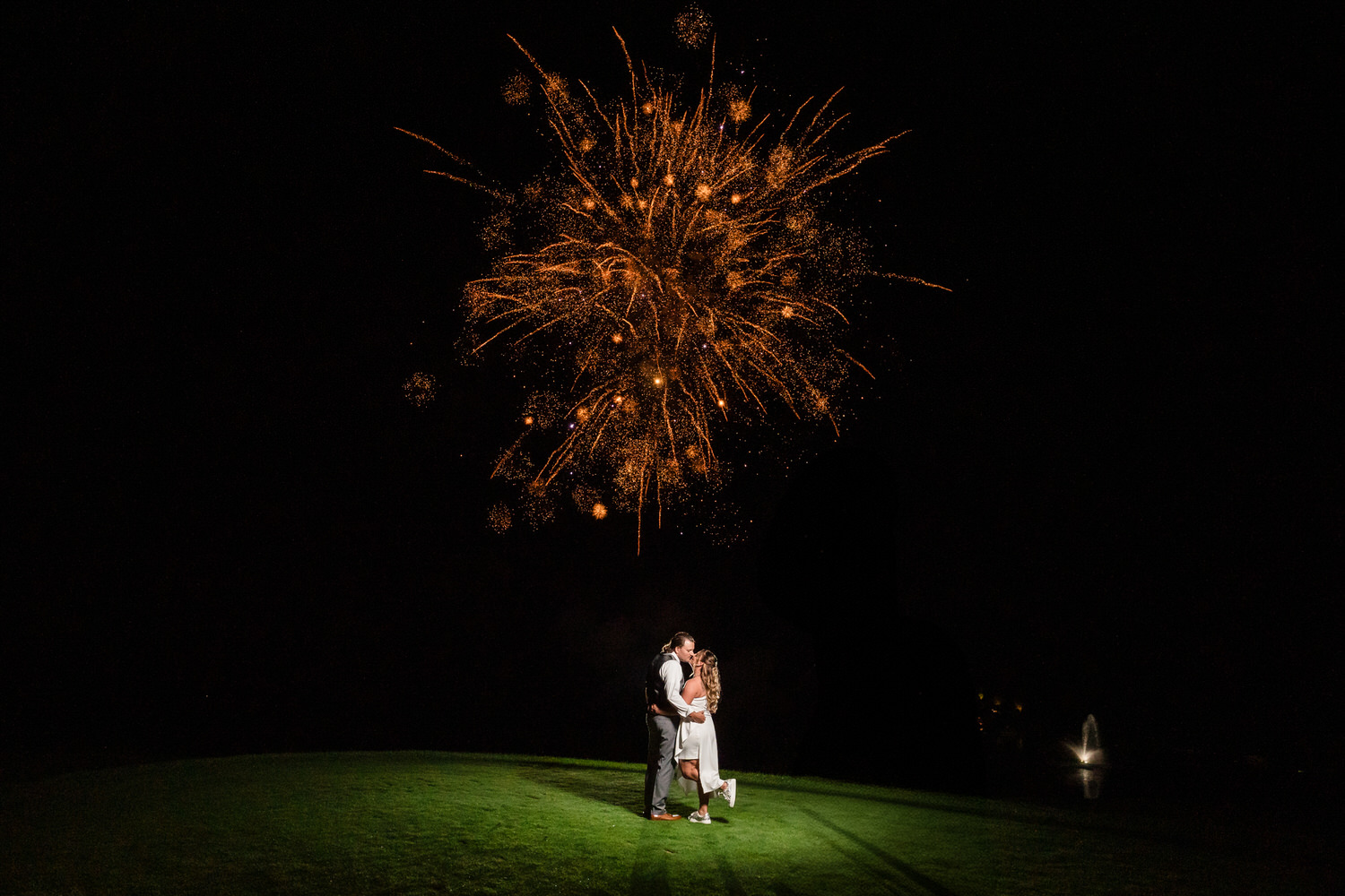 Wide-angle shot of a couple embracing under the golden sparks of a professional wedding fireworks display.