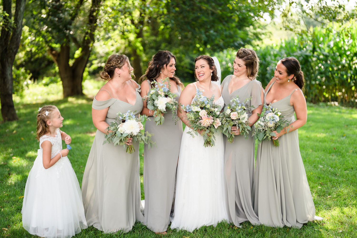 Bride and bridesmaids in sage gowns holding bouquets and smiling in a garden.