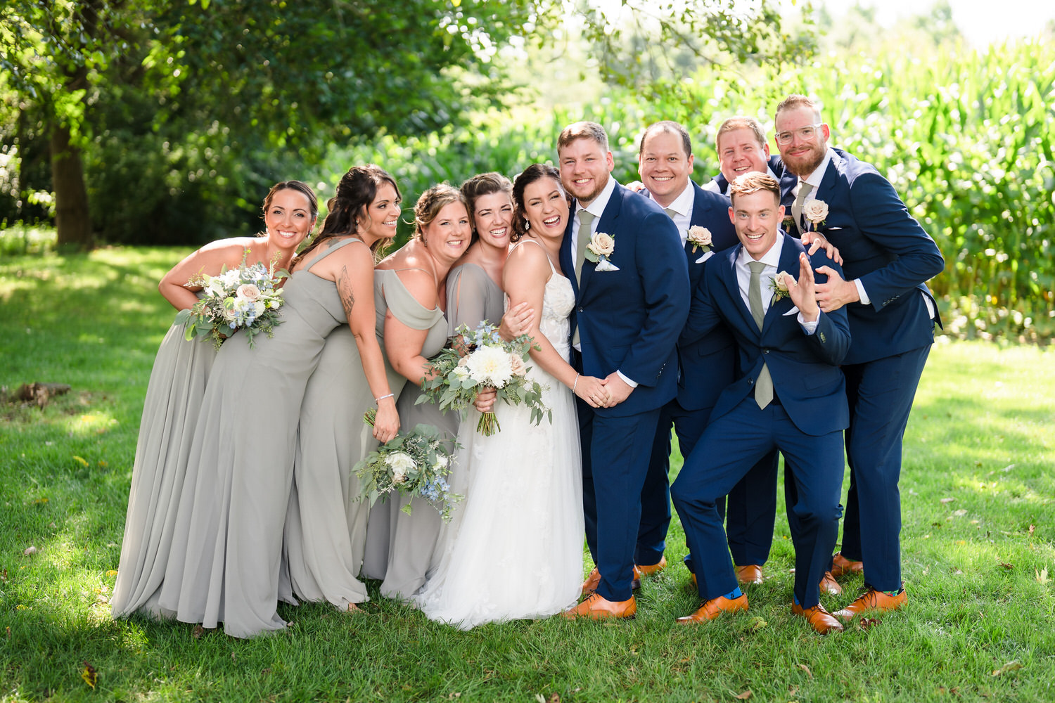 Large wedding party in navy suits and sage dresses posing in a lush green field.