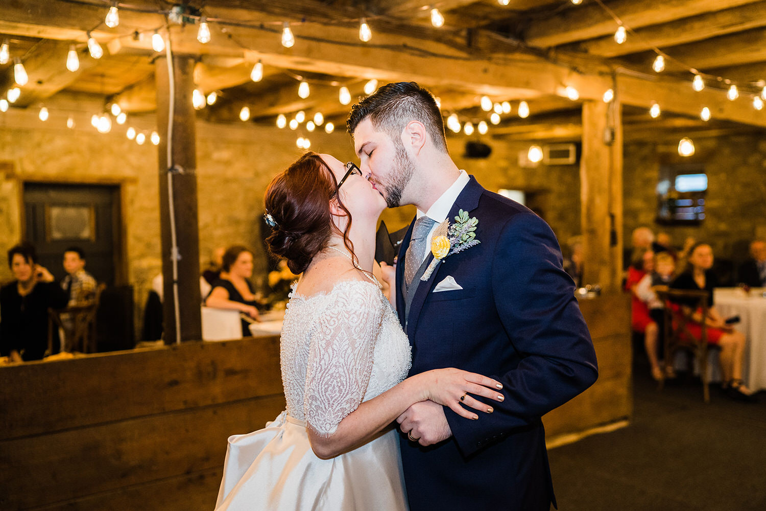 Bride and groom sharing a kiss under string lights in the rustic barn at House and Barn in Emmaus, Lehigh Valley PA.