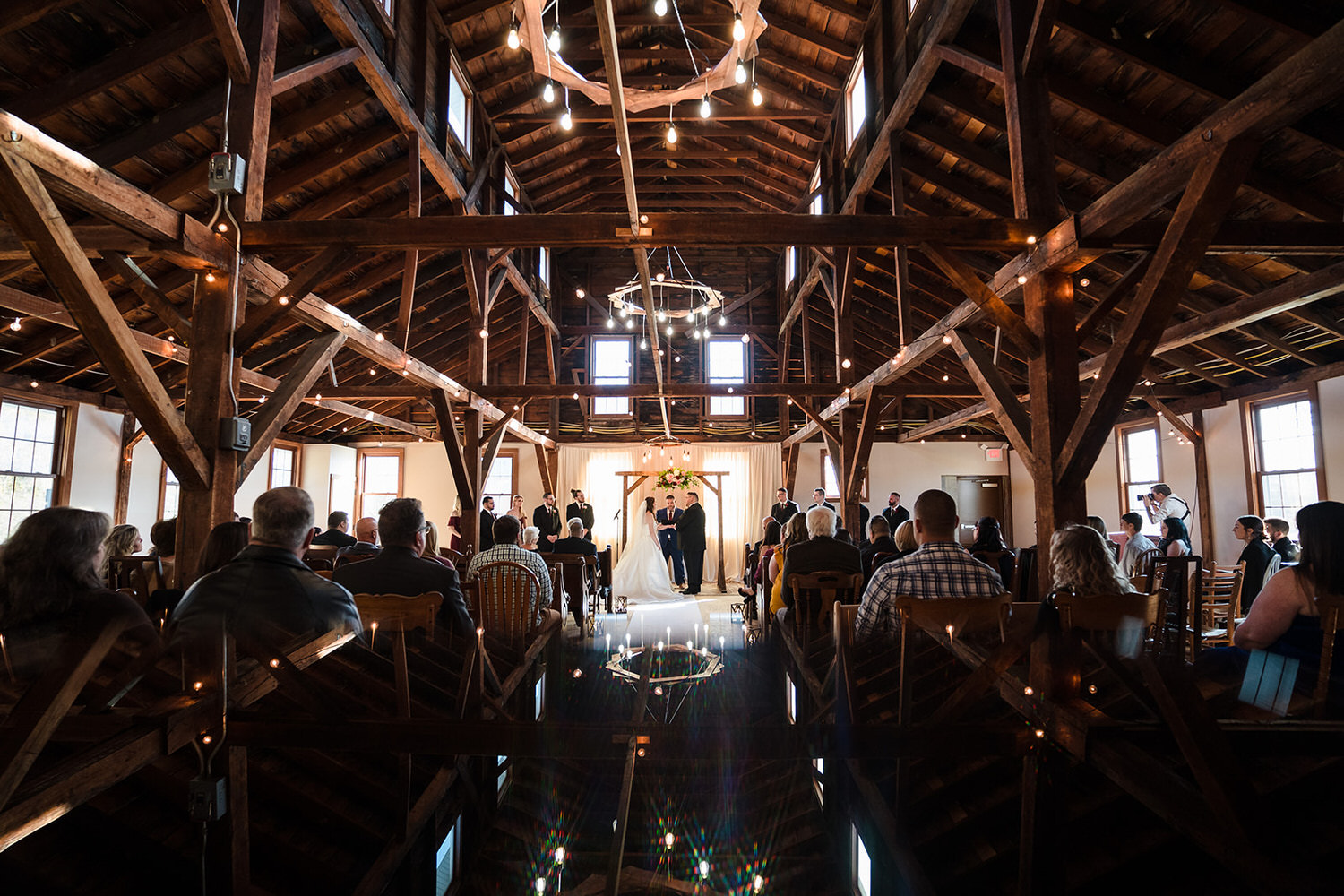 Indoor wedding ceremony featuring a wooden arch and hanging light fixtures in the barn-style event space at Llantrisant Event Venue in Tatamy, PA.
