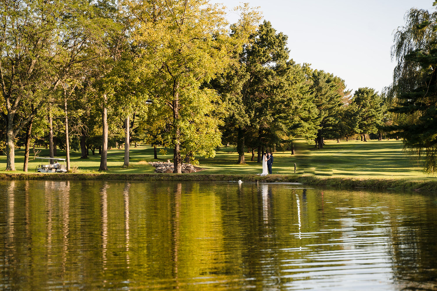 Wedding couple standing by a tranquil pond with lush tree reflections on the golf course at Steel Club in Hellertown, PA.