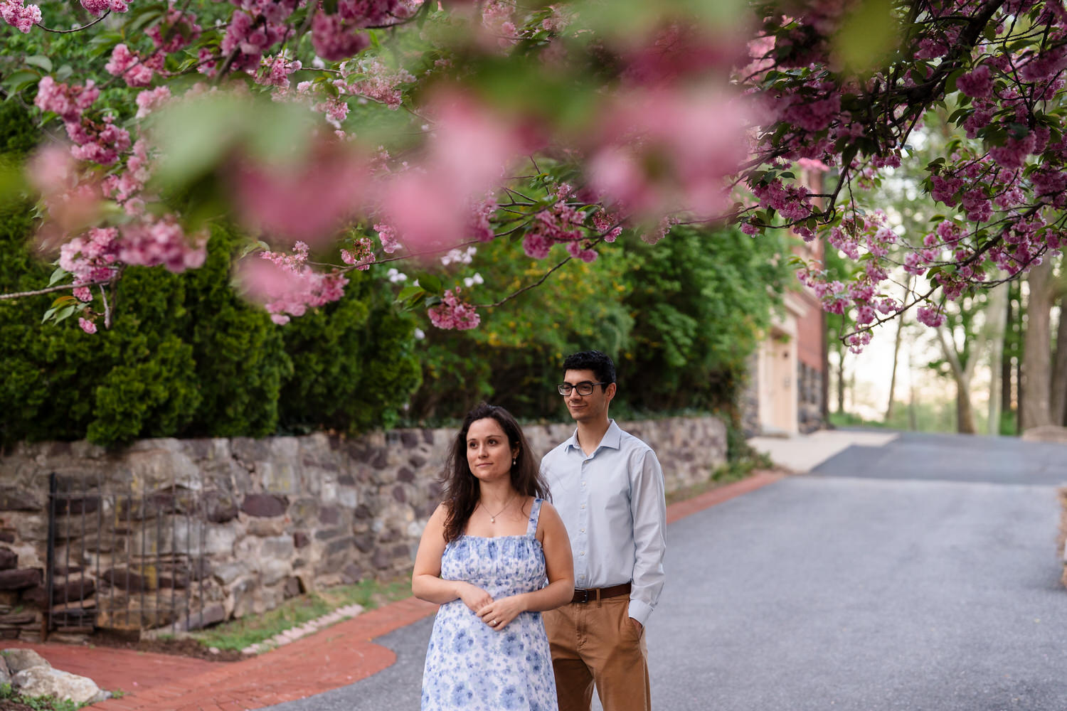 Couple embraces along a brick walkway with cherry blossoms and stone wall
