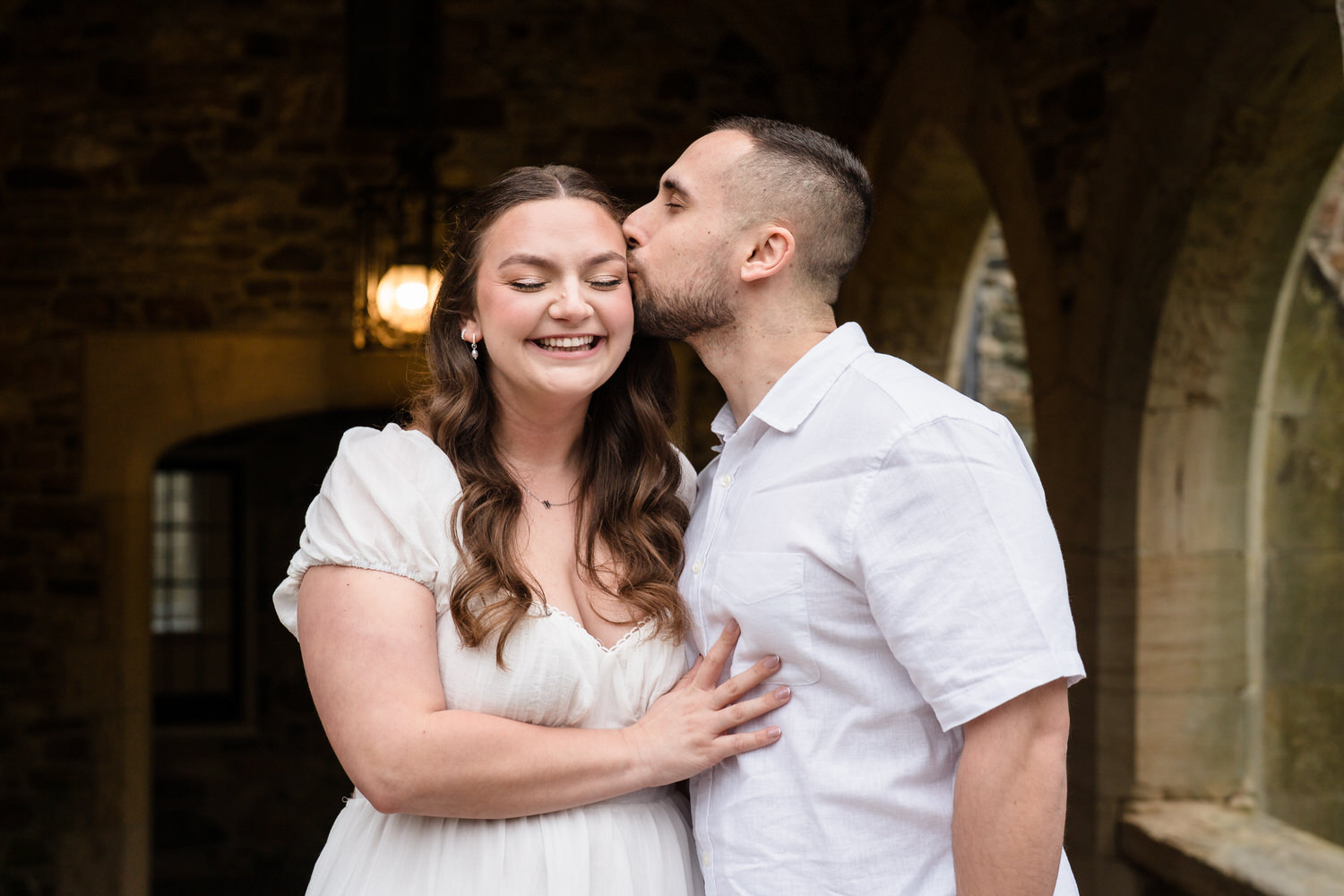 Groom kissing bride’s cheek during warm indoor engagement portrait