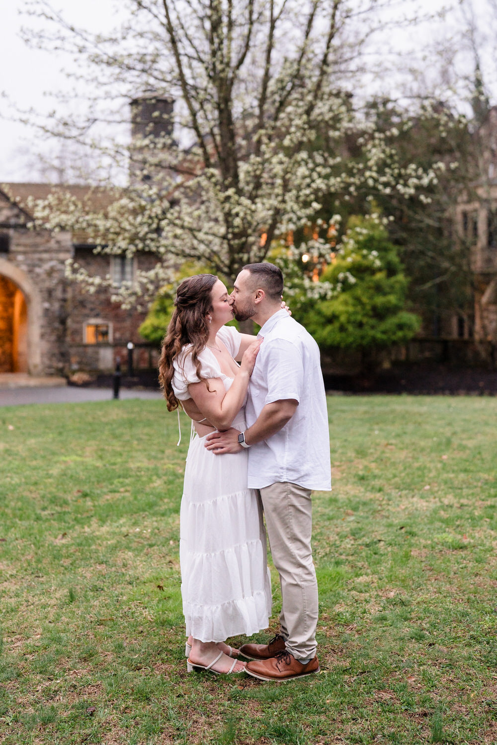 Engaged couple embracing on lawn outside stone estate during spring engagement session
