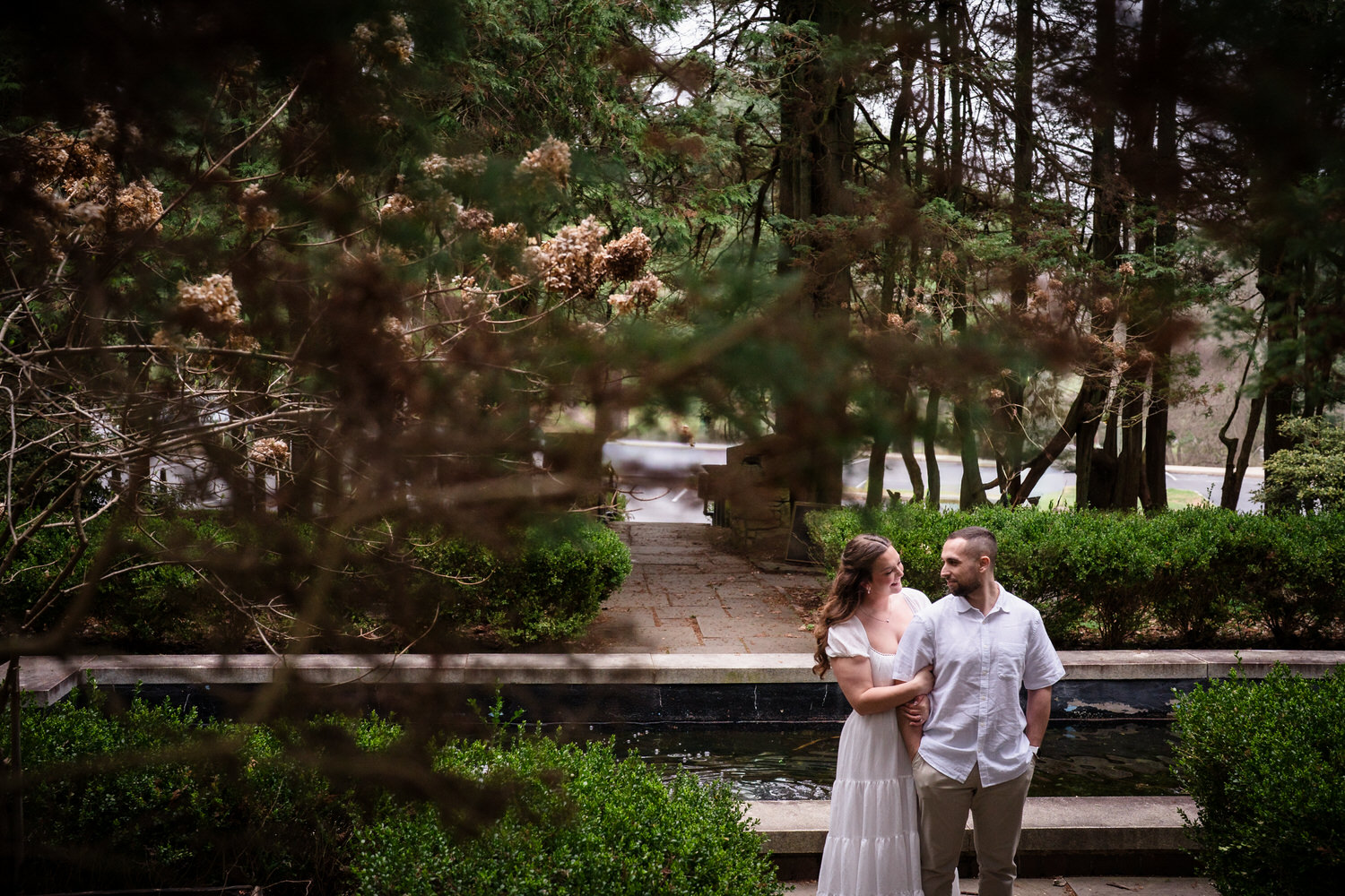 Engagement photo of couple standing near fountain surrounded by greenery