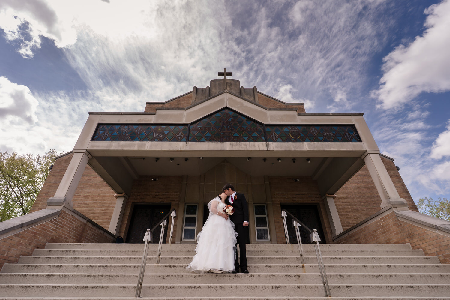 Bride and groom standing on church steps beneath dramatic sky