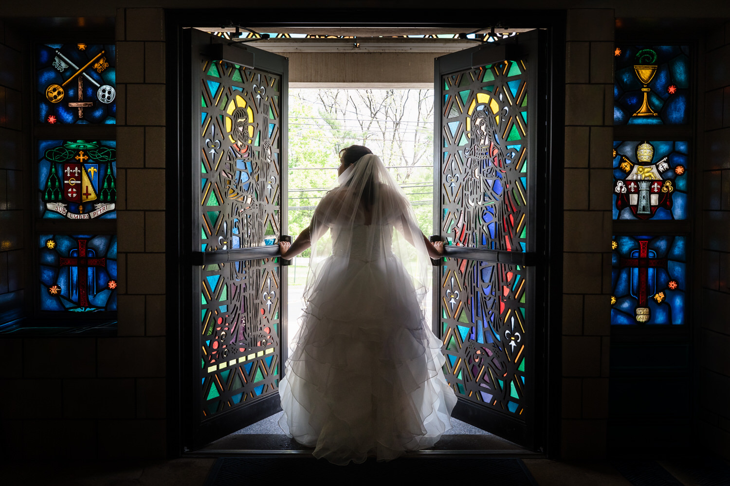 Bride standing in silhouette by colorful stained glass church doors