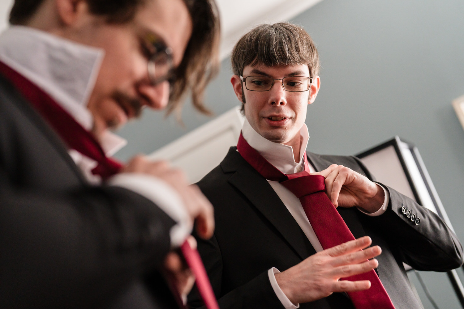 Groom adjusting red tie while groomsman helps before wedding ceremony