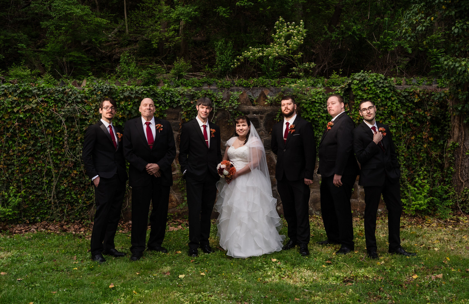 Wedding party portrait with bride, groom, groomsmen, and family standing outdoors in wooded greenery