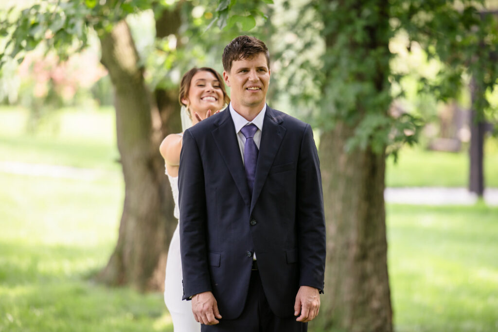 A woman in a white dress stands behind a man in a dark suit outdoors, smiling as she prepares to surprise him. Trees and greenery surround them, creating a bright and joyful atmosphere.