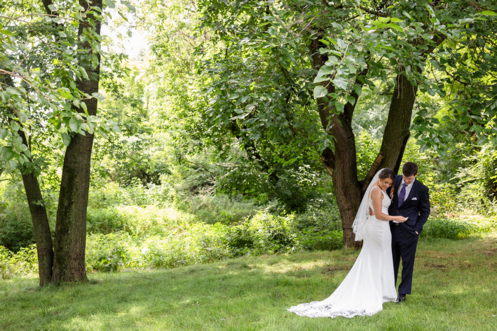 A bride and groom stand together on grass under tall trees, surrounded by lush greenery. The bride wears a white dress and veil, while the groom wears a dark suit. Sunlight filters through the leaves above them.
