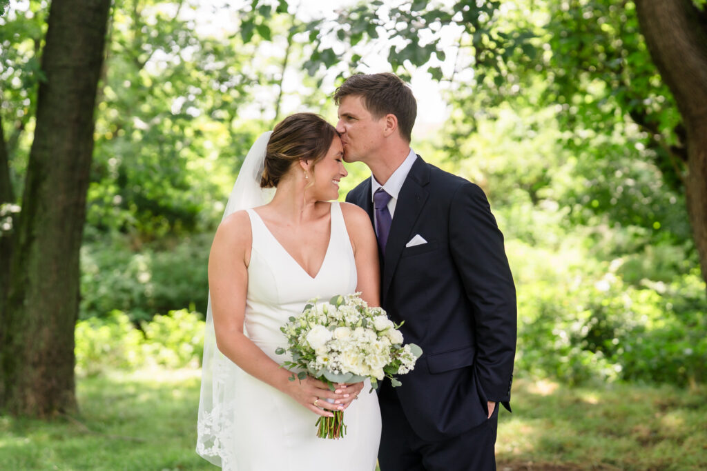 A bride in a white dress holding flowers smiles as a groom in a dark suit kisses her forehead in a lush, green outdoor setting.
