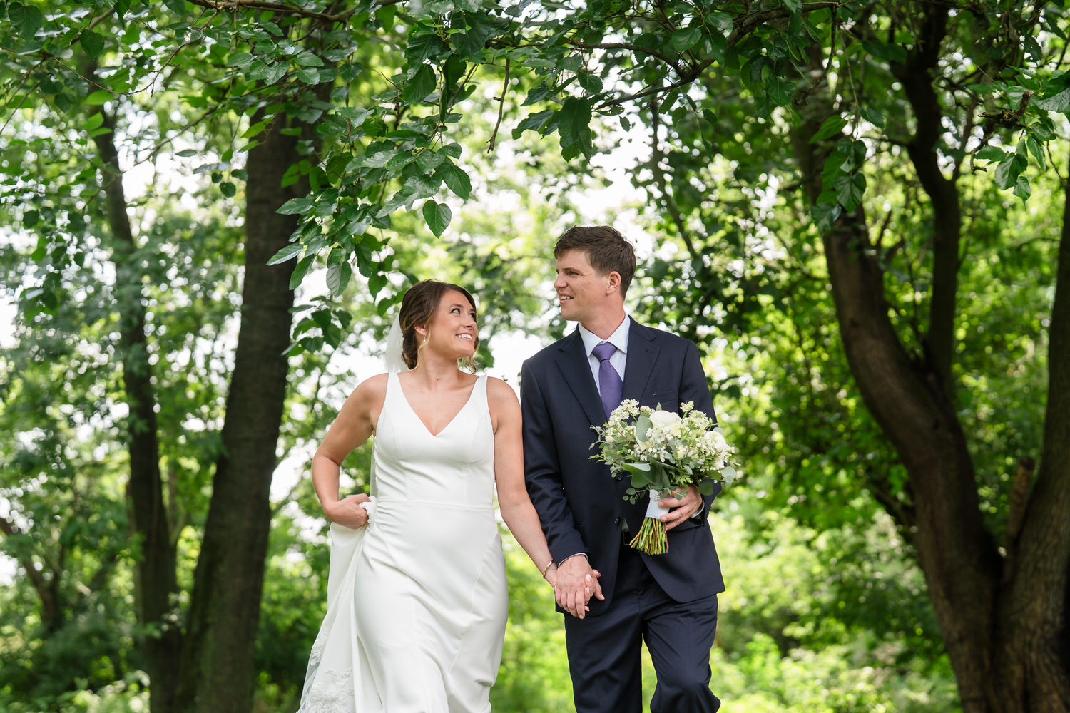 A bride and groom walk hand in hand under green trees. The bride wears a white dress and smiles at the groom, who holds her hand and a bouquet of flowers. Both look happy and are surrounded by lush greenery.