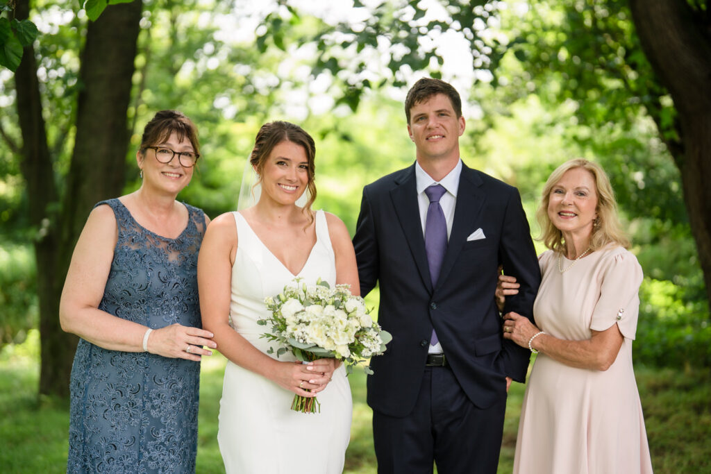 Four people pose outdoors: a bride in a white dress holding flowers, a groom in a dark suit with a tie, and two older women in formal dresses, all smiling in a green, wooded setting.