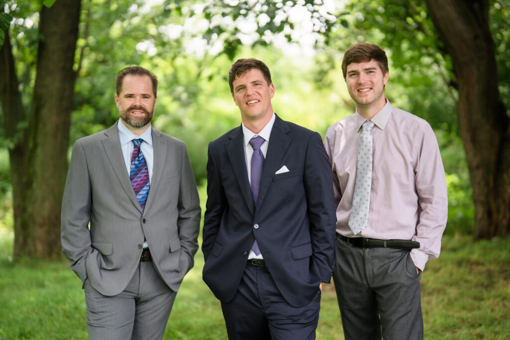 Three men stand outdoors in a grassy, wooded area. All wear dress shirts and ties; two wear suits. They are smiling and have their hands in their pockets. The background is bright and lush with green trees.