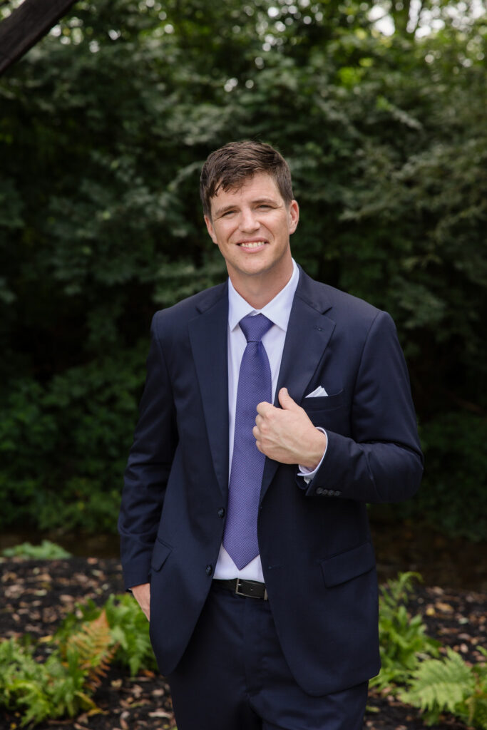 A man in a navy suit and purple tie stands outdoors, smiling at the camera with one hand adjusting his jacket. Green shrubs and plants are in the background.