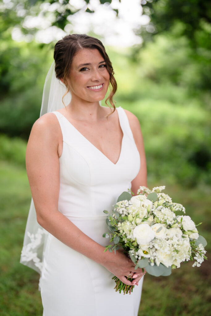 A bride in a sleeveless white gown and veil smiles while holding a bouquet of white flowers and greenery, standing outdoors with a blurred green background.