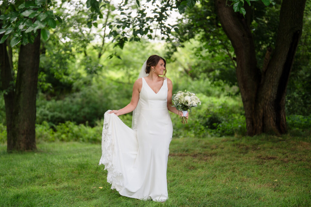 A bride in a white dress holds a bouquet and lifts her train while standing on green grass in a lush, wooded area.