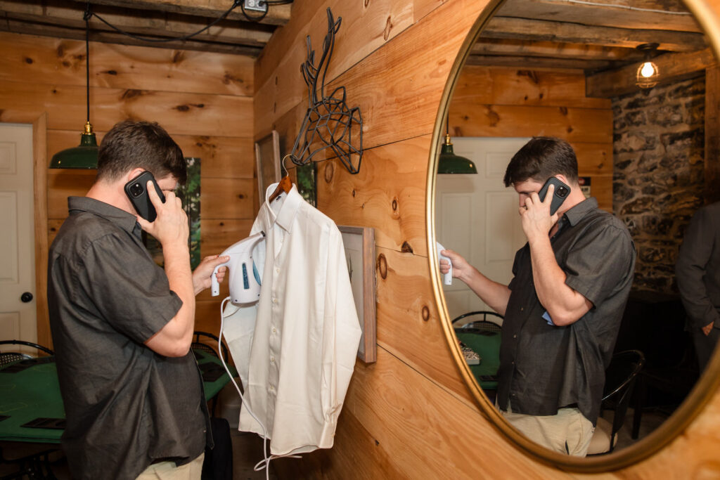 A man talks on the phone while using a steamer on a white shirt, standing in a wooden room. His reflection is visible in a large round mirror on the wall.