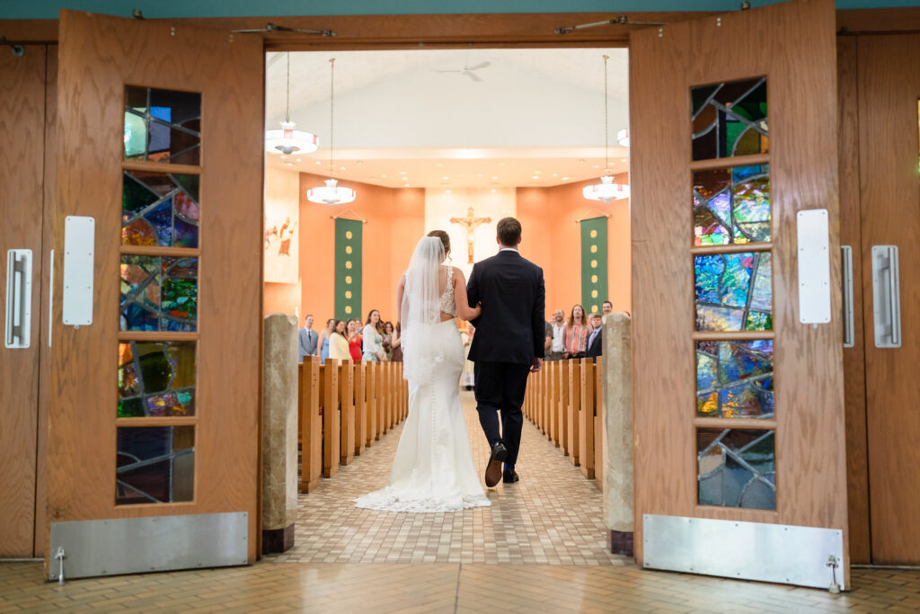 A bride and groom walk down the aisle together in a church, with wedding guests seated on either side and colorful stained glass panels on the open doors.
