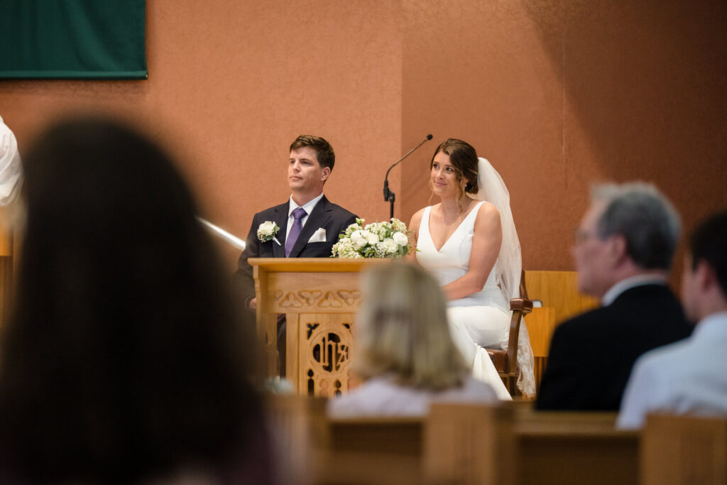 A bride and groom sit together at the front of a church during their wedding ceremony, with floral arrangements nearby and guests seated in the foreground.