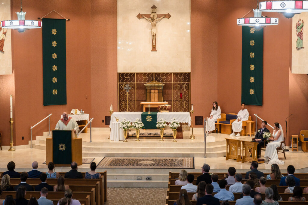 A wedding ceremony in a church shows a priest at the lectern, a couple seated with bouquets, attendants nearby, and guests watching from pews. The altar and religious symbols are visible in the background.
