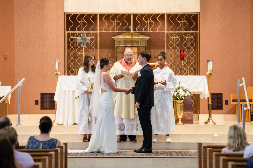 A bride and groom stand holding hands at the altar during a church wedding ceremony, with a priest and two altar servers in white robes standing behind them.