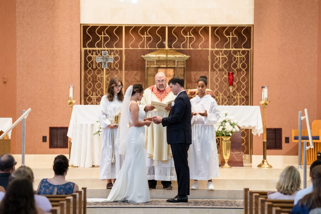 A bride and groom exchange rings during a church wedding ceremony, with a priest and two altar servers standing behind them at the altar. Guests are seated in the foreground, watching the scene.