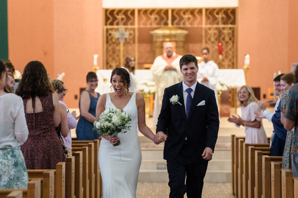 A joyful bride and groom walk down the aisle, holding hands, as guests smile and clap in a church decorated for their wedding ceremony.