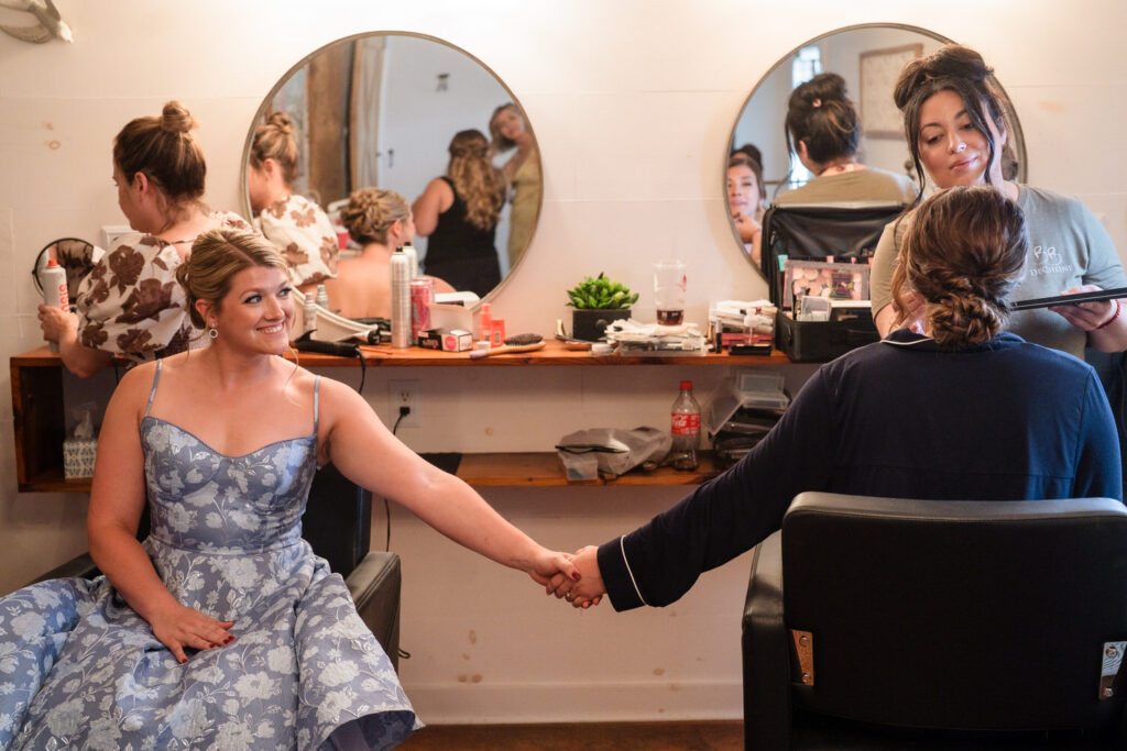 Two women in a salon hold hands; one wears a blue dress and smiles, while the other sits getting makeup applied. Two more women and mirrors are visible in the background. The scene is warm and friendly.