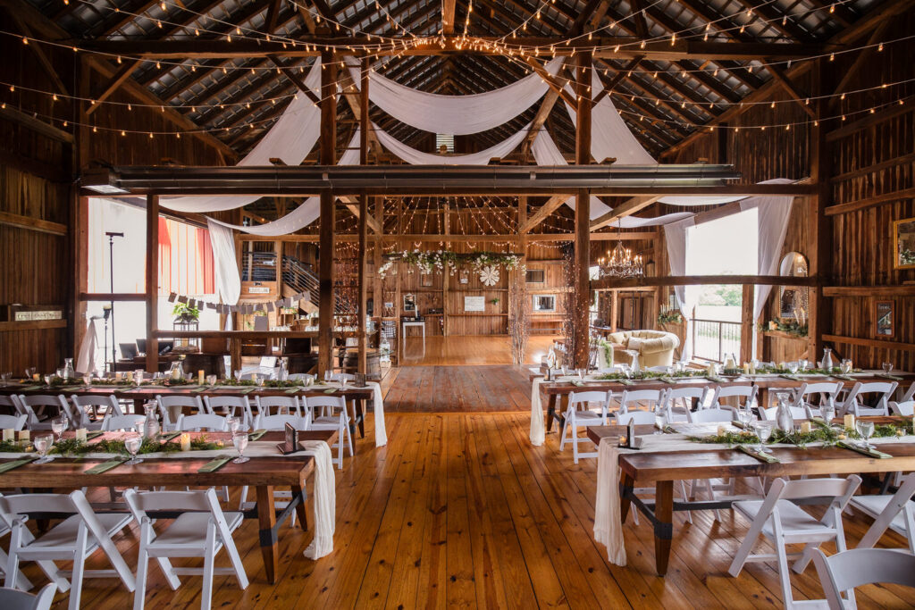 Rustic barn wedding venue with wooden floors, long decorated tables with white chairs, string lights, draped fabric on the ceiling, and a bright, open, welcoming atmosphere.