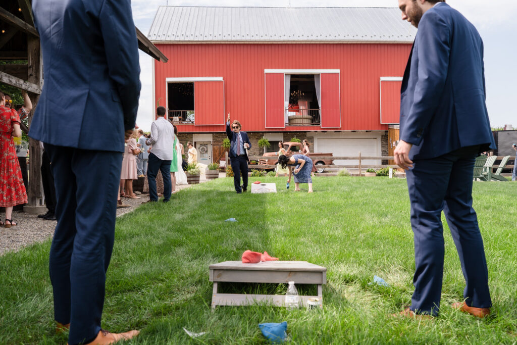 People in formal attire play cornhole on grass beside a red barn. A man throws a bean bag, while others watch and socialize in the background. The setting appears to be a festive outdoor gathering or wedding.