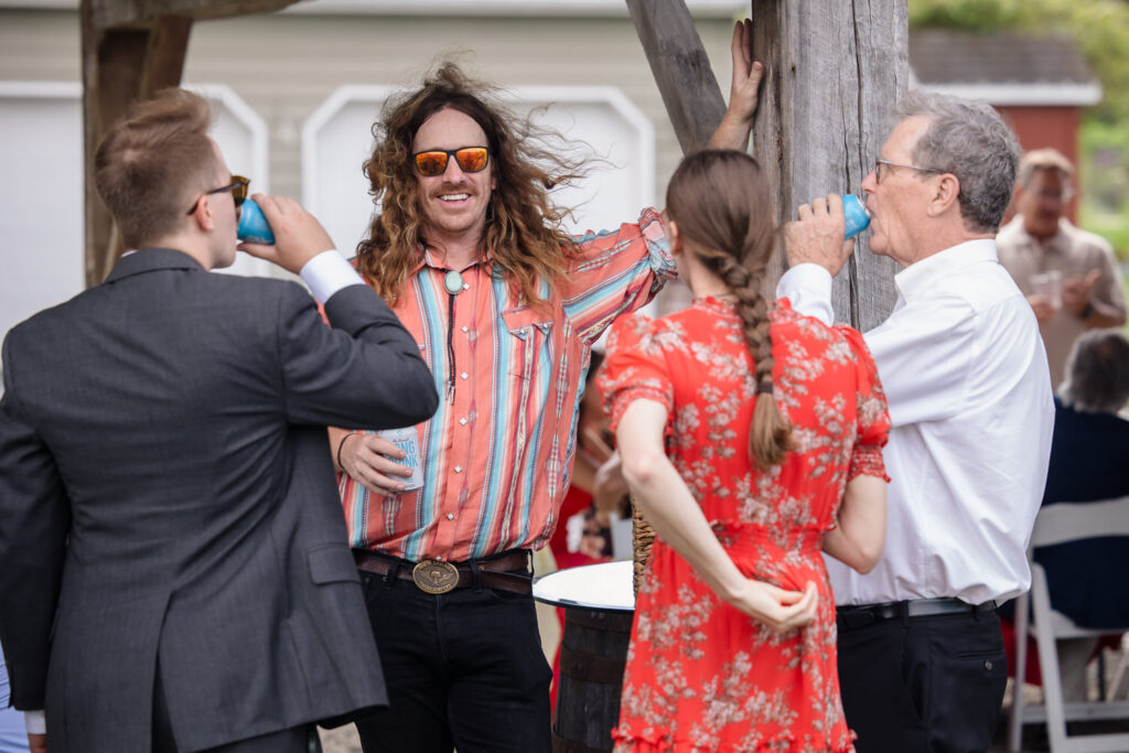 Four adults stand in a circle outdoors, talking and drinking from cans. One man with long hair and sunglasses leans on a wooden post, smiling. Another man wears a suit, and a woman in a red dress has her back to the camera.