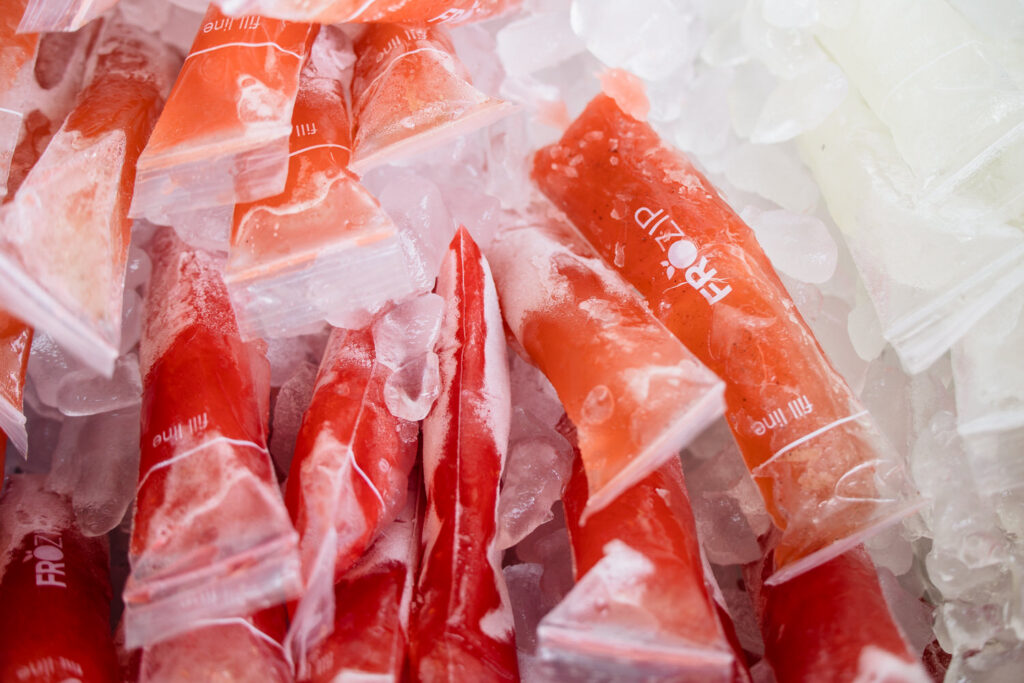 Red and white frozen ice pops in clear plastic sleeves are piled on ice. The text FROZIP appears on some of the sleeves. The pops display frost and condensation from the cold.