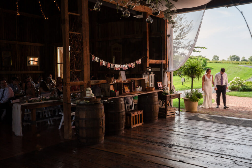 A bride and groom enter a rustic barn wedding venue, holding hands. Guests are seated inside. Sunlight streams through the open door, highlighting greenery and trees outside.