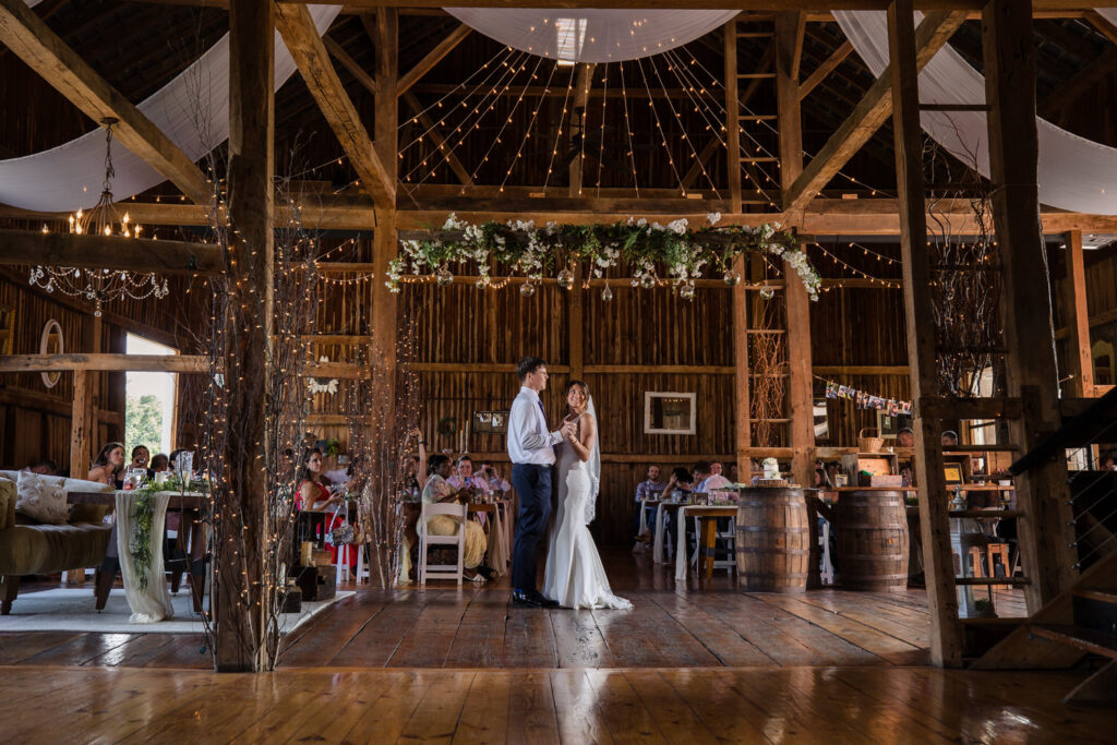 A bride and groom share their first dance in a rustic barn decorated with string lights and flowers, while guests seated at tables watch them.