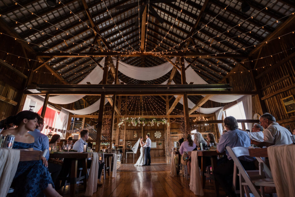 A couple dances in the center of a rustic barn decorated with string lights and white drapes, while seated guests watch and enjoy the celebration.