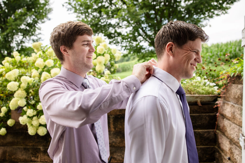 Two men stand outside near blooming flowers; one man in a dress shirt helps the other adjust his tie. Both are smiling, and they appear to be preparing for a formal event.