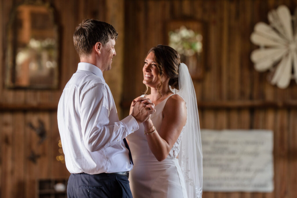 A bride and groom smile and hold hands while dancing together in a rustic wooden venue, with sunlight softly illuminating their faces.
