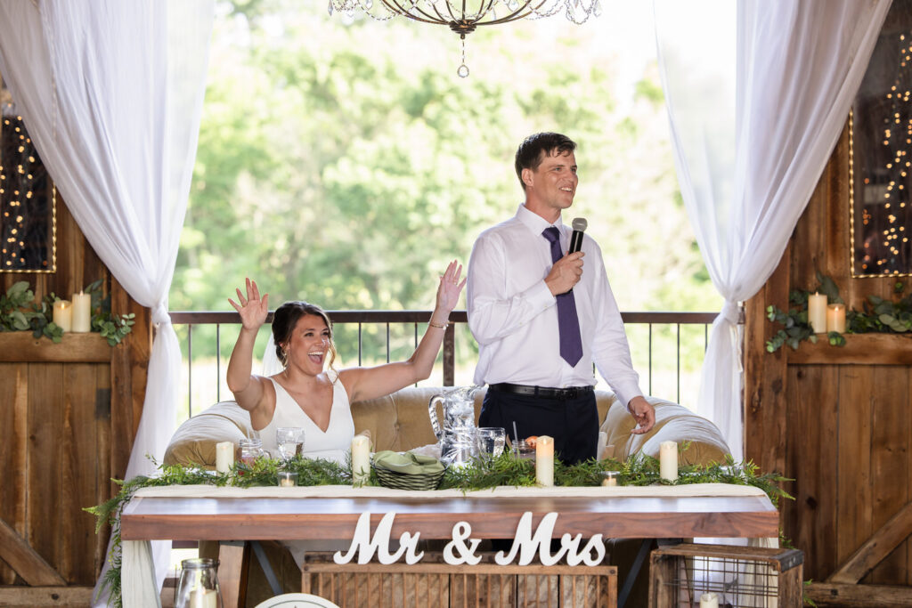A bride sits smiling with raised arms while the groom stands beside her holding a microphone. They are at a decorated table with a Mr & Mrs sign, in a rustic venue with greenery and candles.
