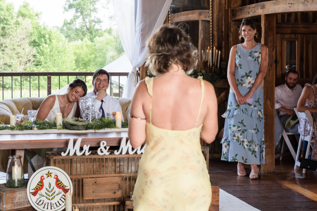 A woman in a yellow dress gives a speech at a rustic wedding. The bride and groom sit at a decorated table, smiling. Another woman in blue stands nearby, watching. Guests sit in the background.