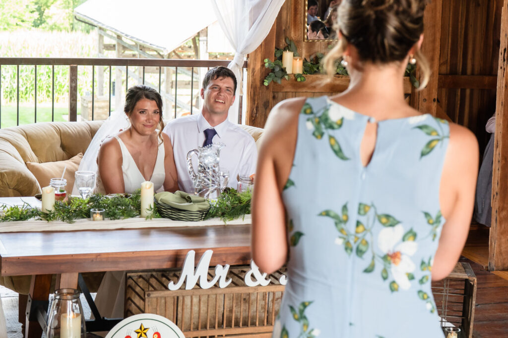 A bride and groom sit smiling at a decorated table while a woman in a blue floral dress stands facing them, giving a speech in a rustic, wooden venue.