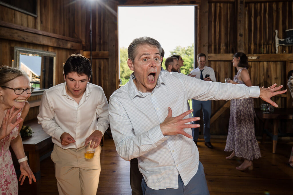 A man in a light shirt poses with a surprised expression and outstretched arms at a lively indoor gathering, while others laugh and mingle in the background.